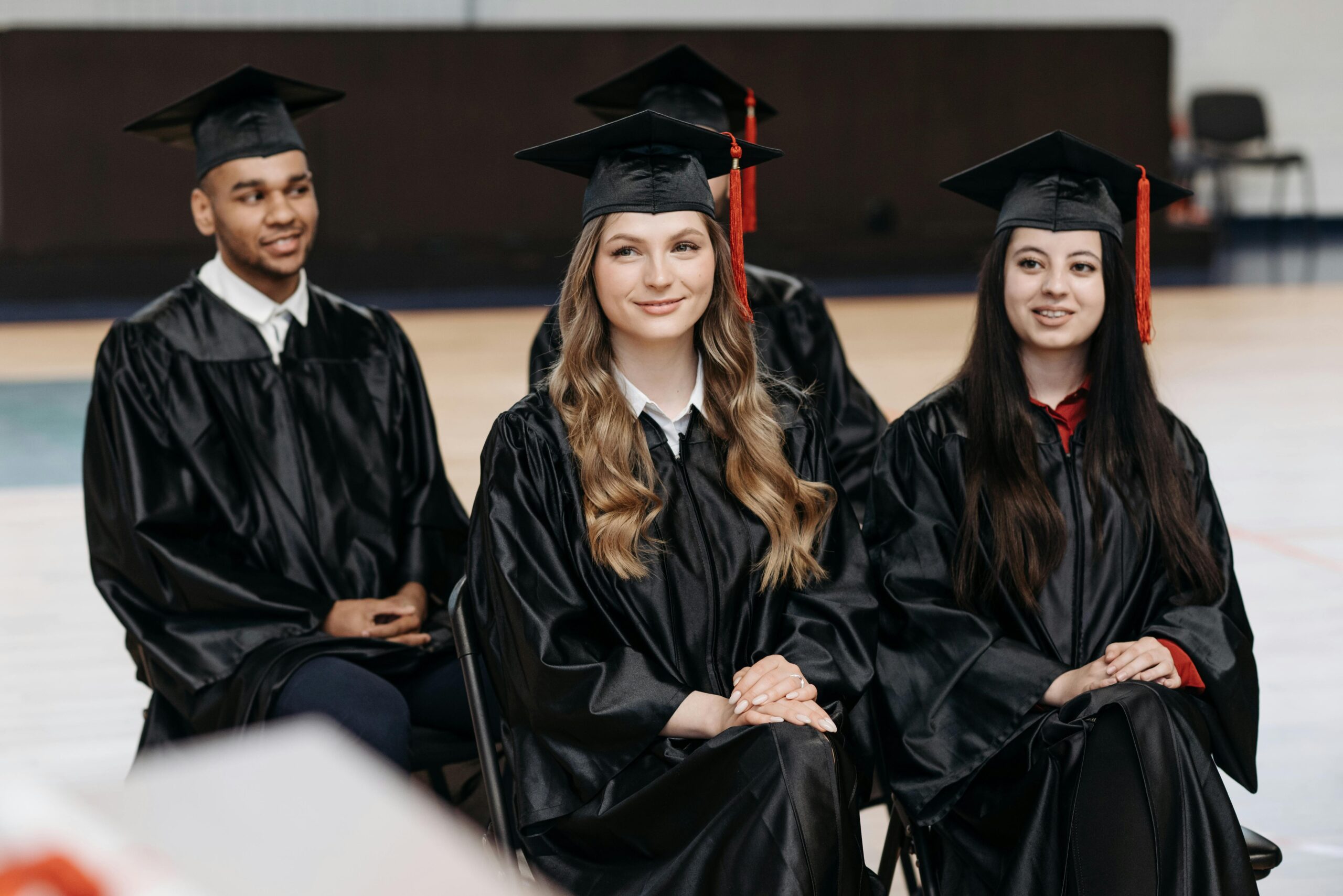 Four graduates in caps and gowns seated during a graduation ceremony, smiling.